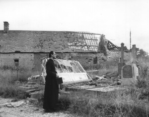 Parish priest at St. Jores, France, Father Le Courtois, eyes battle damage to the church and graveyard - July 7, 1944. (U.S. Army Signal Corps Photo, Courtesy National Archives)